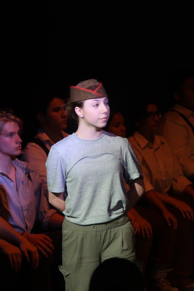  Actor in grey shirt and wearing a soldier hat speaking as others are seated behind and looking on. 
