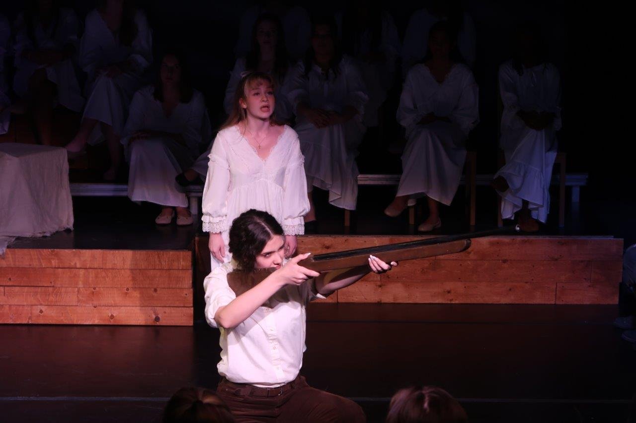  Actor in front crouched and aiming a rifle to their left, while another actor behind them in a white dress looks on. 