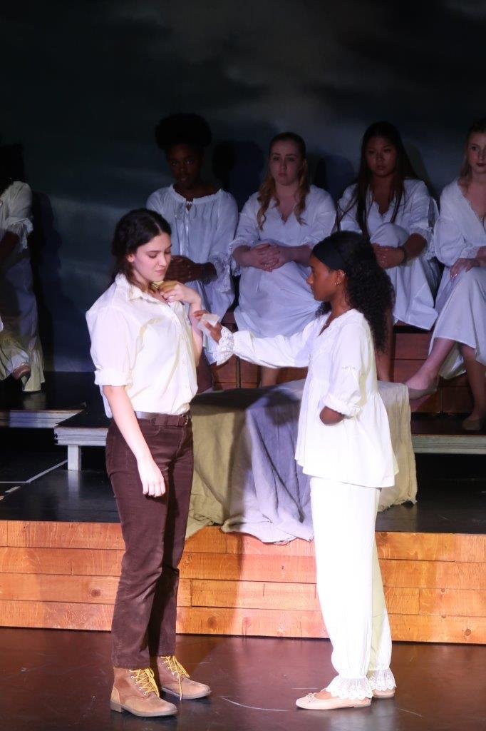  Actor on left in white shirt and brown pants touches their collarbone while the actor on right in all white puts out their right hand towards the other while holding a note. 