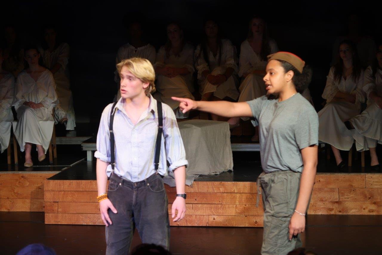  Actor on right in grey shirt and soldier’s cap points to their right, as the actor on left in white shirt and suspenders looks in the pointed direction. 