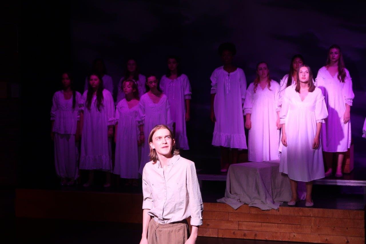  Acttor in foreground in white shirt spotlighted along with another actor standing in the background surrounded by others.  All looking in the same direction to their left. 