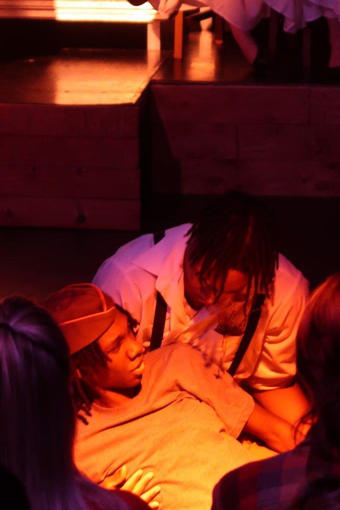  Actor in soldier’s cap being held by another in white shirt and suspenders. 