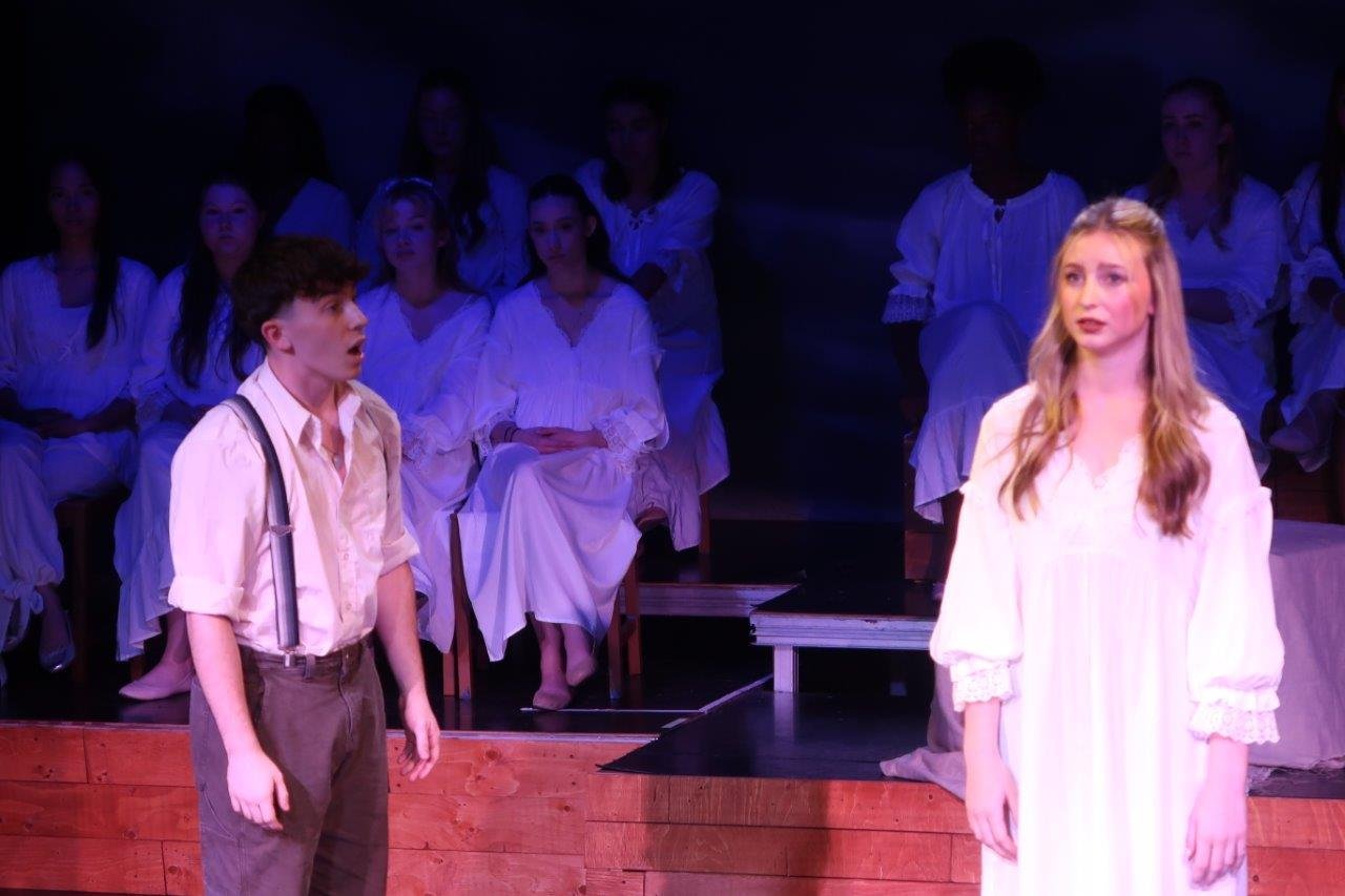  Actor on left in white shirt and suspenders speaking to their left as actor on right in white dress looks forward, as others in the background look on. 