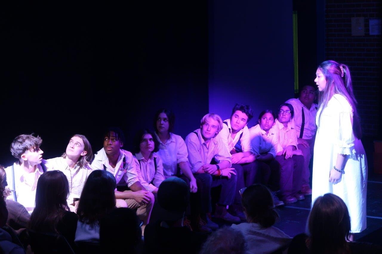  Actor dressed in white on the right speaking to an actor seated on the left.  To the right of the seated actor, others look up and away. 