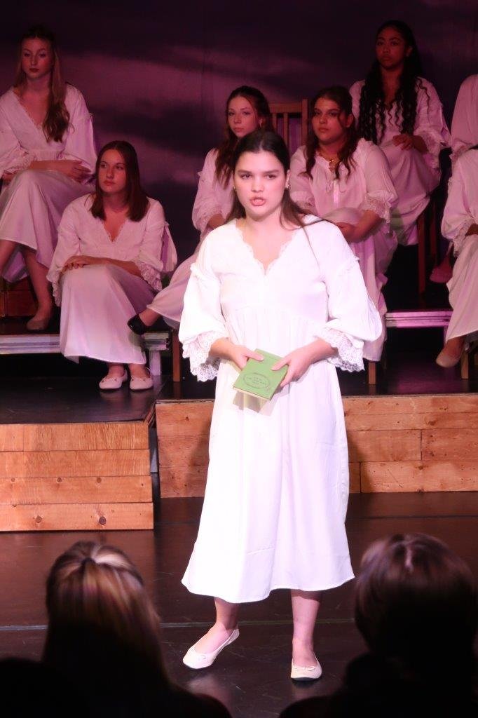  Actor in white dress speaking as others looks on from behind. 