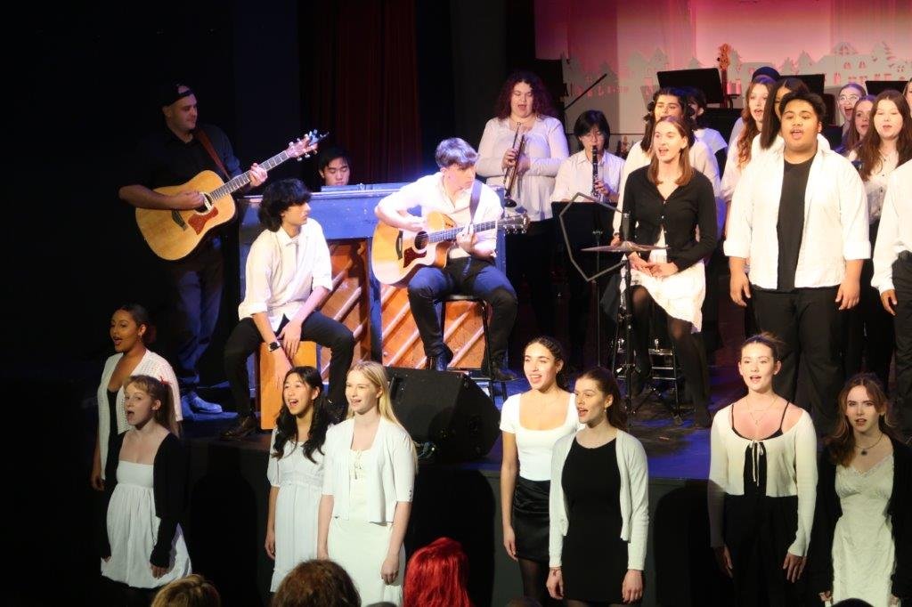  Group of singers in white singing, with accompanists playing guitar, beatbox, and instruments to the left and behind. 