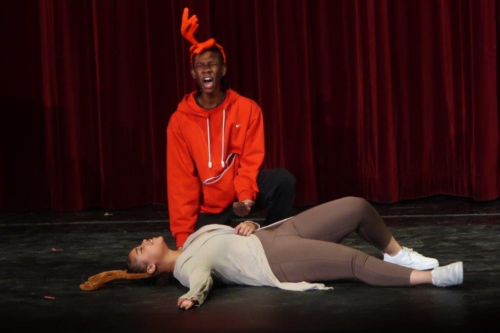  Student dressed in red with red reindeer antlers screaming as another student in brown with brown reindeer antlers lies on the floor in front of them. 