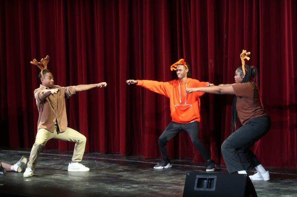  Actor in red with red reindeer antlers pointing at two actors in brown with brown reindeer antlers  on either side of one another. 