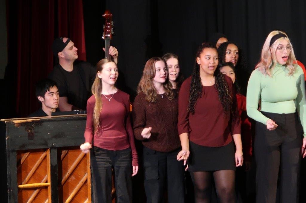  Group of singers singing against a black background with piano and cello accompanists to the left. 