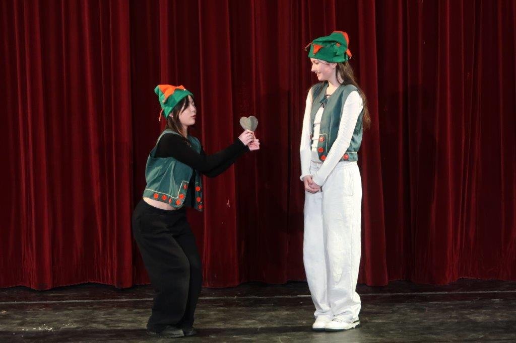  Actor on left wearing an elf hat giving a heart to an actor wearing white, an elf vest, and an elf hat on the right. 