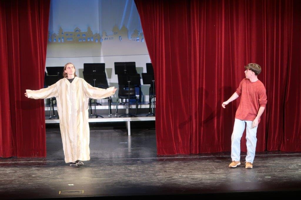  Curtains parting to reveal an actor dressed in a white robe, while on the right an actor wearing red looks back in surprise. 