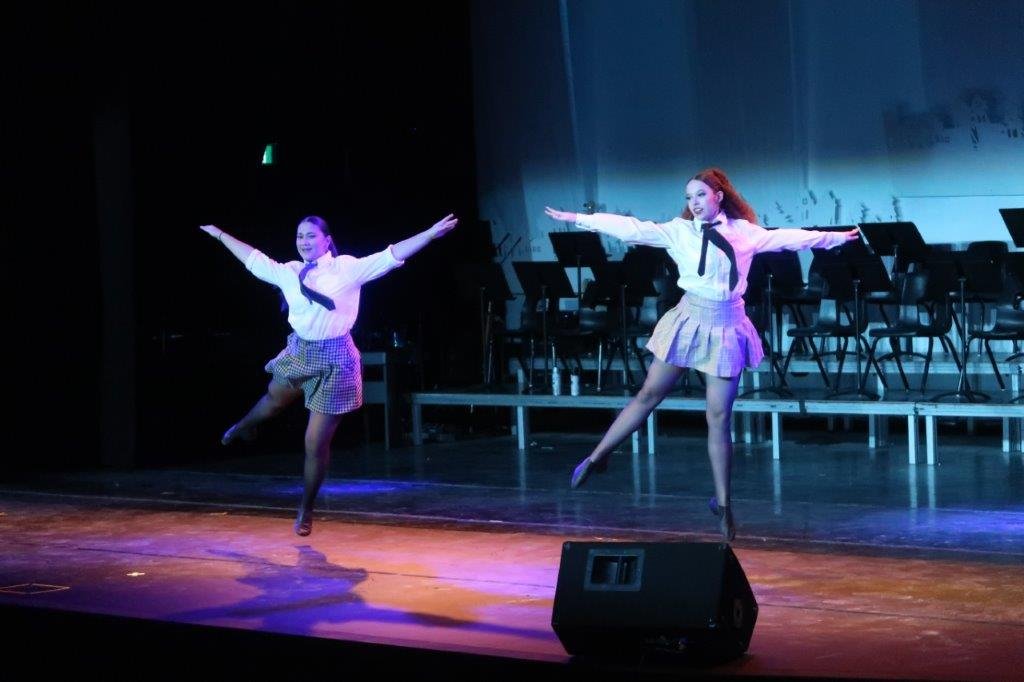  Two dancers wearing white shirts, ties, and skirts are jumping on stage. 