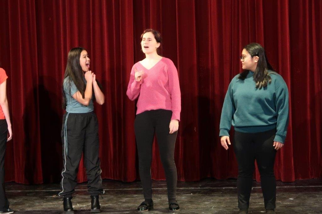  On the left, an actor in blue top and black pants looks excited as they look upon an actor in pink speaking in the centre.  On the right, a narrator in green looks on the scene. 