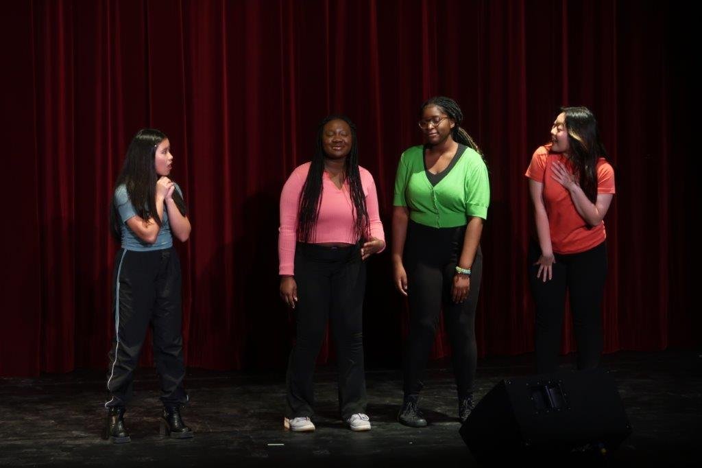  On the left, actor in blue top and black paints looks disappointed as three actors in pink, green, and orange look on. 