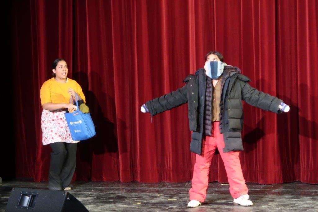  Actor on left dressed in yellow shirt and pink apron is pulling out clothes from a bag towards actor on right wearing a number of layers of tops and jacket with face covered by a large scarf. 