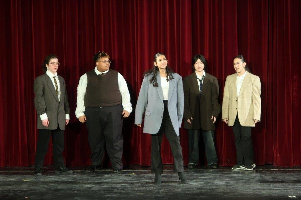  Group of actors, some dressed in suit jackets, against a red curtain. 