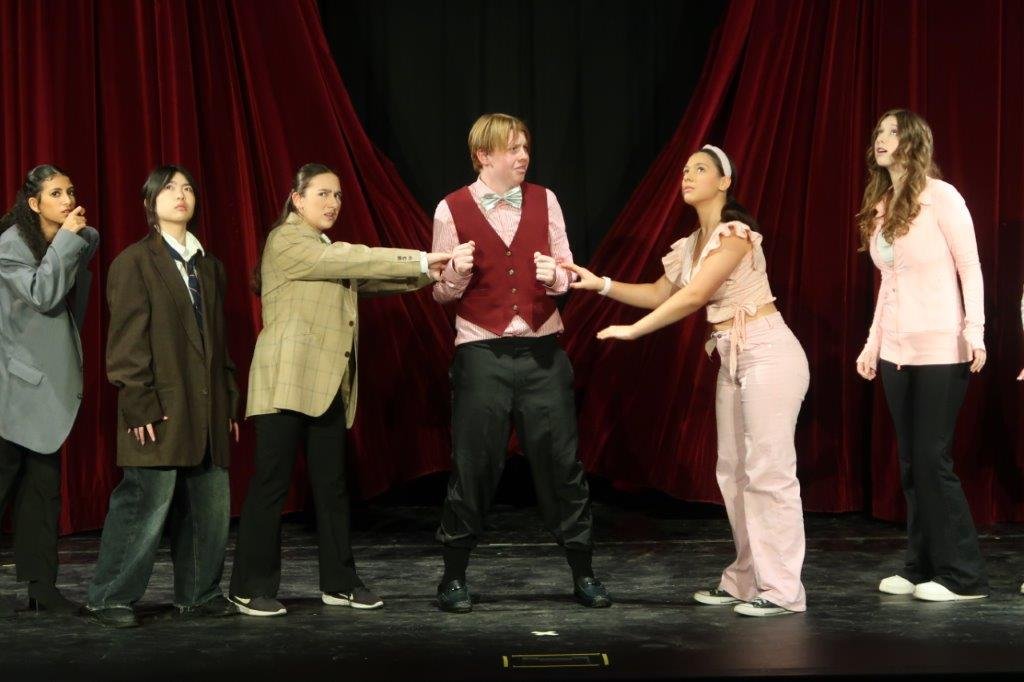  Actor in pink button shirt, red vest, and blue bowtie being held on left arm by an actor wearing pink, and being held on right arm by actor in a brown suit jacket. 