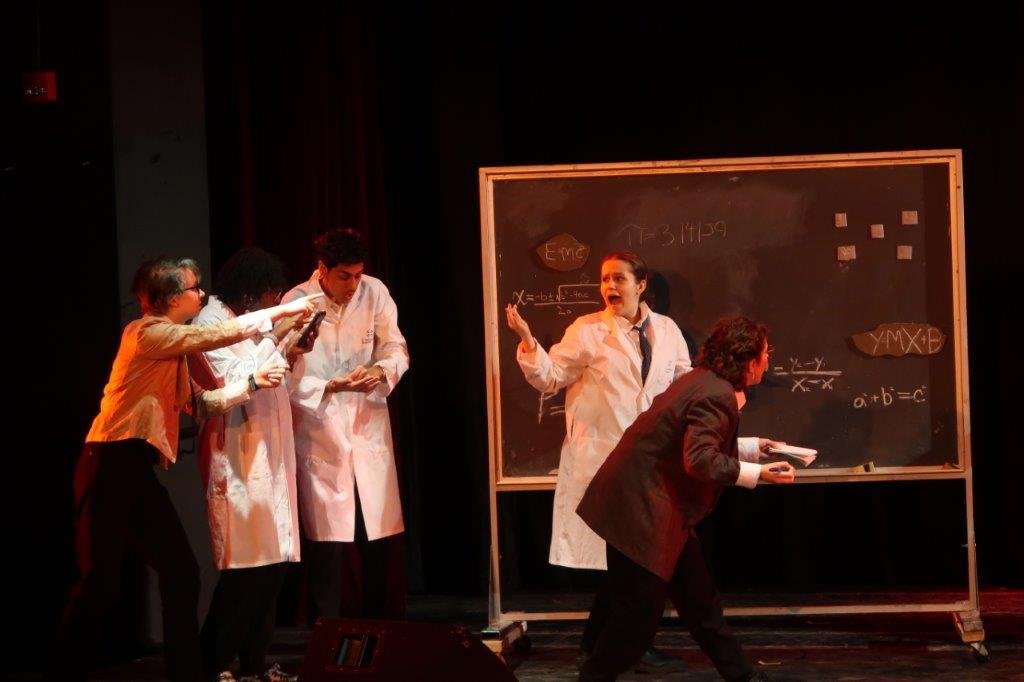  Actors dressed in lab coats at a chalkboard with formulae written on the board. 