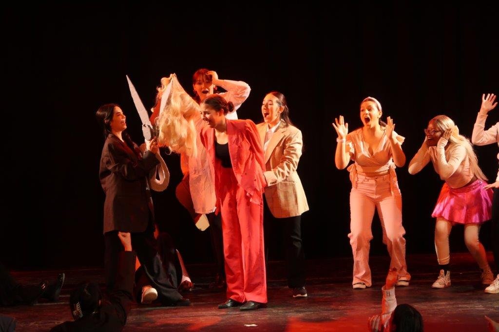  Actors in pink on the right look on screaming as actor in centre with pink suit and pants has their hair cut off by an actor on the left wearing a brown suit jacket and holding a giant pair of scirros. 