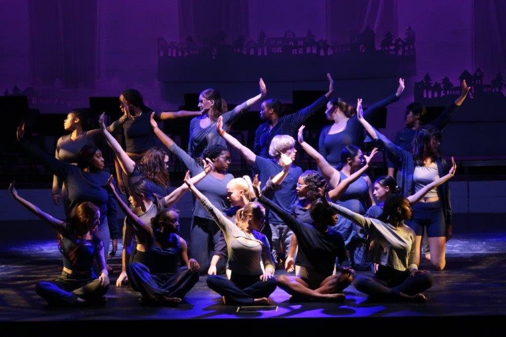  Dancers kneeling in the foreground facing and pointing their arms away in rows. 