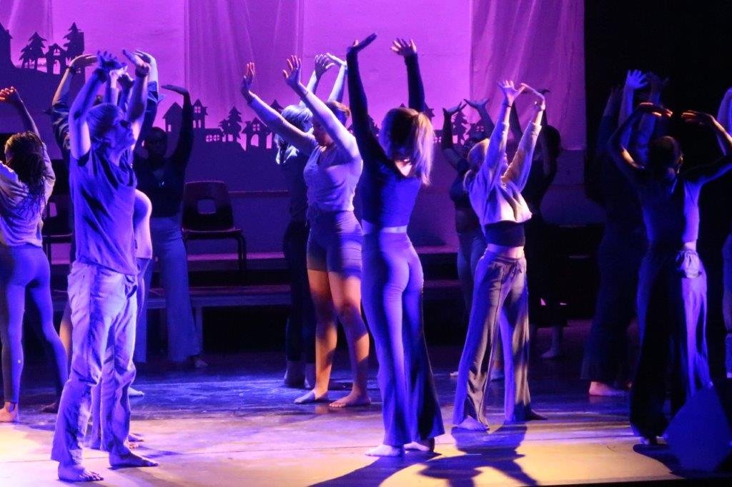  Dancers standing with their arms outstretched above them are castr in a purple-blueish light. 