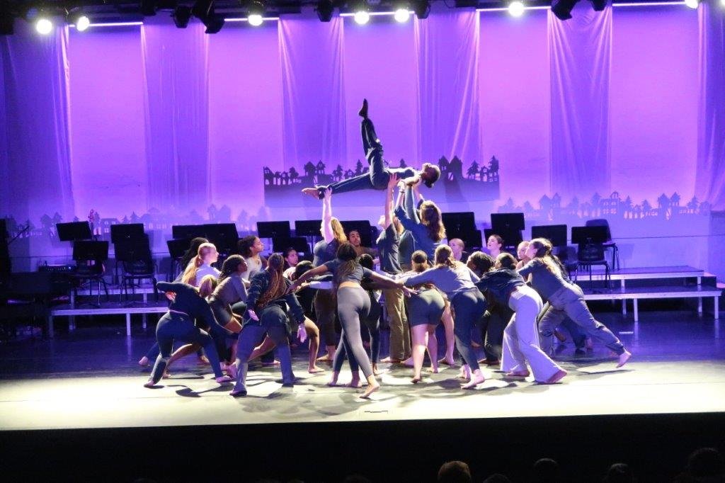  Group of dancers congregating towards the centre, with some dancers holding up one dancer in the air with one leg parallel to the ground and the other leg lifted straight up. 