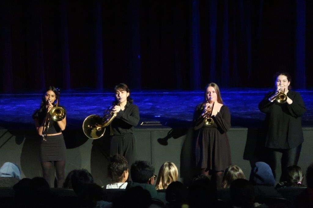  Four musicians in black playing their brass instruments at the front of the stage. 