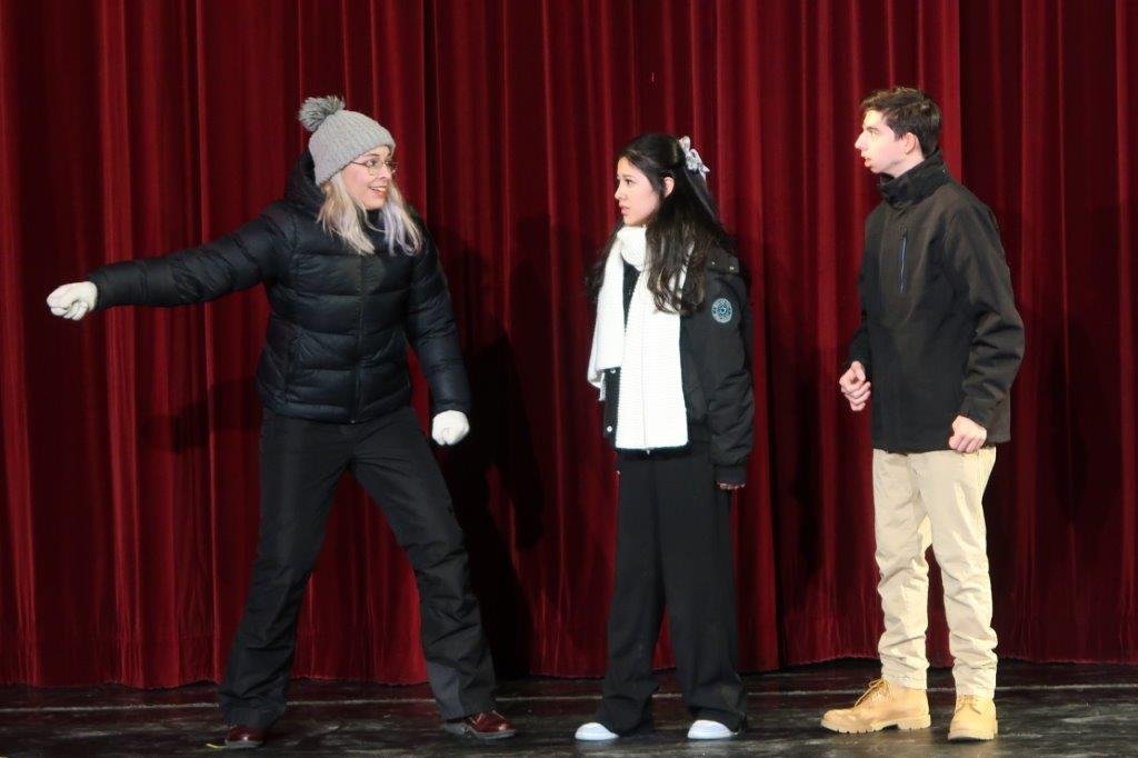  Three actors dressed in winter jackets.  The actor on the left has their right arm outstretched and speaking to the other two looking at them. 