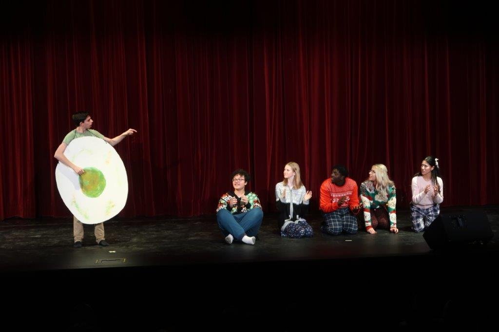  Five actors sitting on the stage to the right while an actor stands on the left dressed as an egg with a green yolk. 