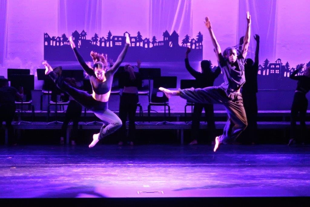  Two dancers jumping with right feet outstretched and arms up in the air bathed in purple stage light. 