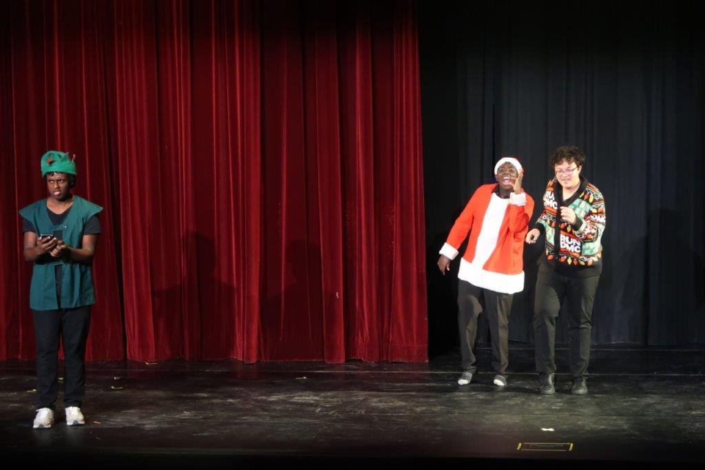  An actor on the far left dressed in a green elf outfit looks at his phone as two actors dance on the right.  One actor is dressed in a Santa outfit and the other is dressed in a Christmas sweater. 