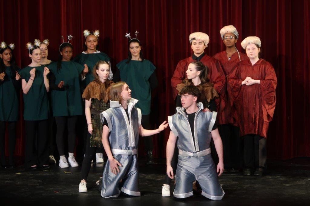  Two actors in the foreground are dressed in silver and kneeling.  Directly behind them are two actors dressed in brown.  In the foreground, there are two groups of actors; the left group is dressed in green while the right group is dressed in red ro