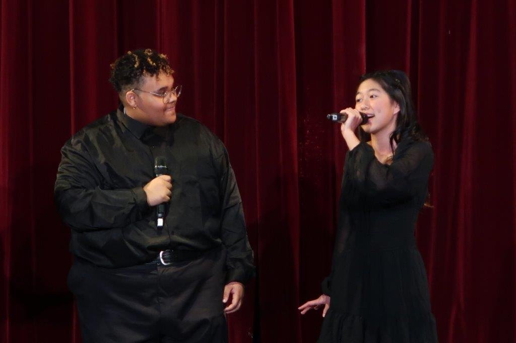  Two singers dressed in black holding microphones against a red backdrop. 