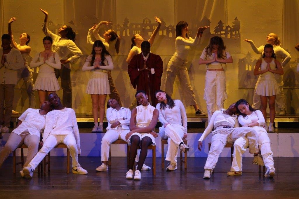  Dancers dressed in white seated in the foreground appearing asleep while other dancers behind them are paused in poses. 