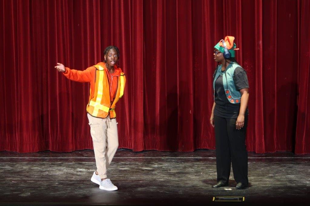  Actor on left dressed in orange and yellow work shirt gesturing as actor on right dressed in black with green elf vest and hat looks on. 