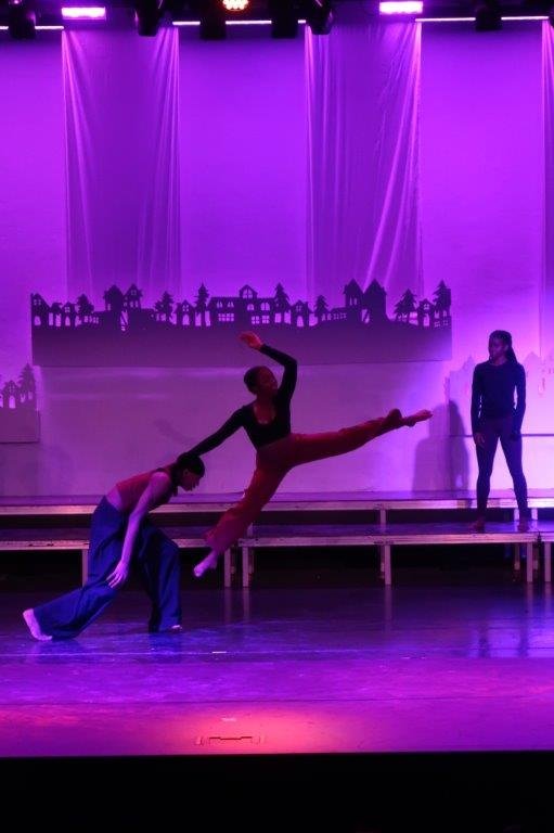  3 dancers; the one of the left is half-bent supporting the middle dancer who has their legs split in mid-jump, while the third dancer on the right looks on in the background. 