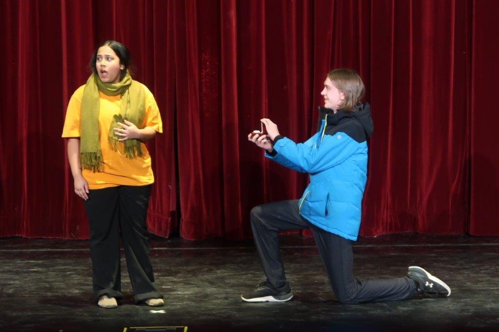  Actor on left  dressed in yellow shirt and green scarflooks off screen as actor on right in blue jacket is on bended knee holding a proposal ring out towards the first actor. 