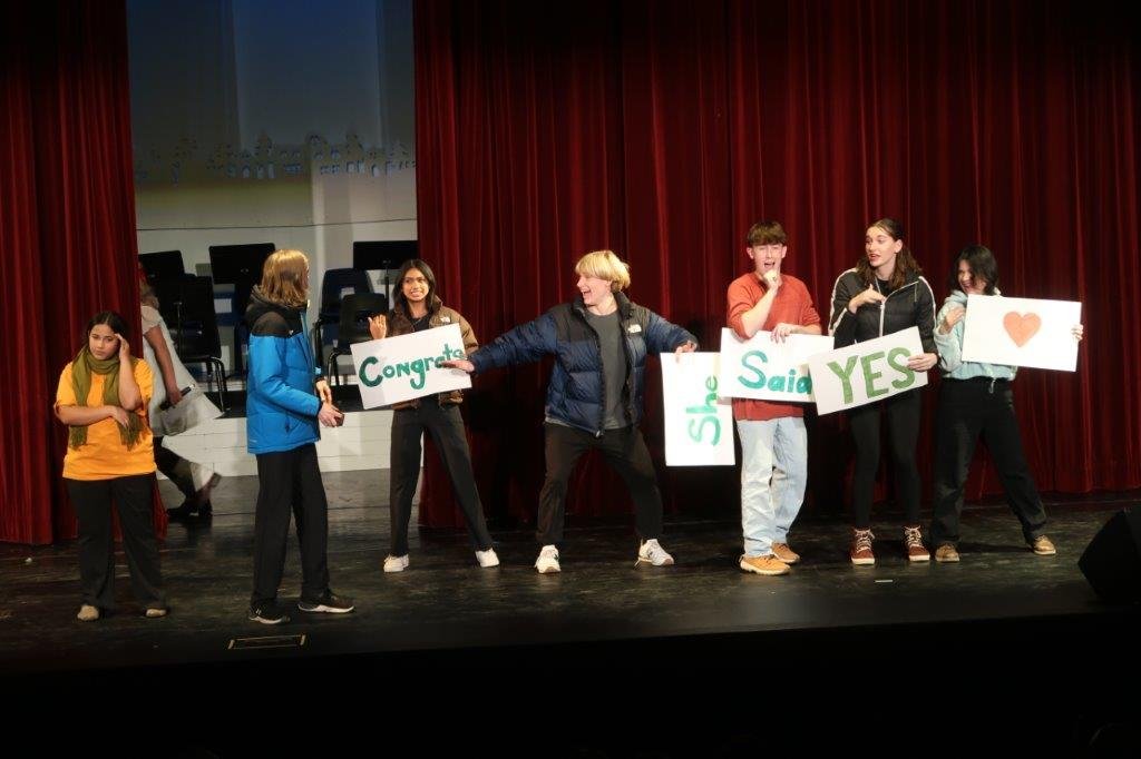  Actors on left wearing yellow and blue look right as five actors holding a sign each with the words “Congrats” “She” “Said” “Yes” “&lt;heart&gt;” 