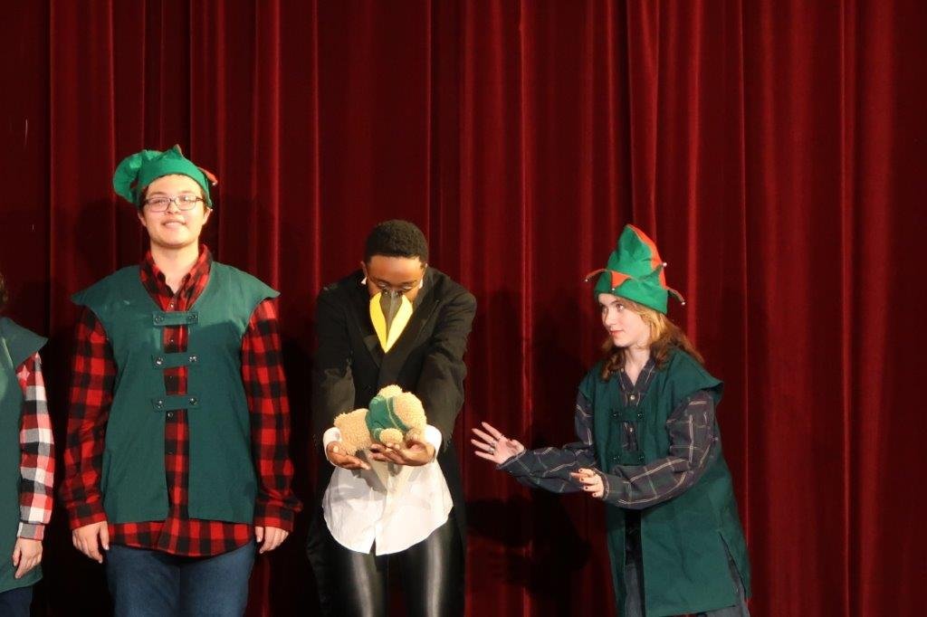  On left, actor in plaid sweater elf vest and elf hat stands straight.  In the middle, an actor with a beak holds a plush bear.  On the right, an actor in elf vest and elf hat waits to be handed the plush bear. 