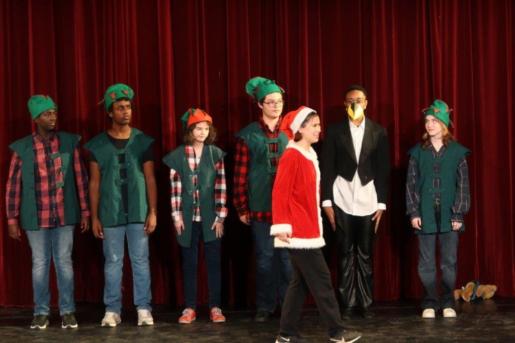  A line of actors dressed as elves in vests and hats in the rear, with one actor dressed in a tuxedo coat and beak second from right, as an actor dressed as Santa Claus walks in front of them. 