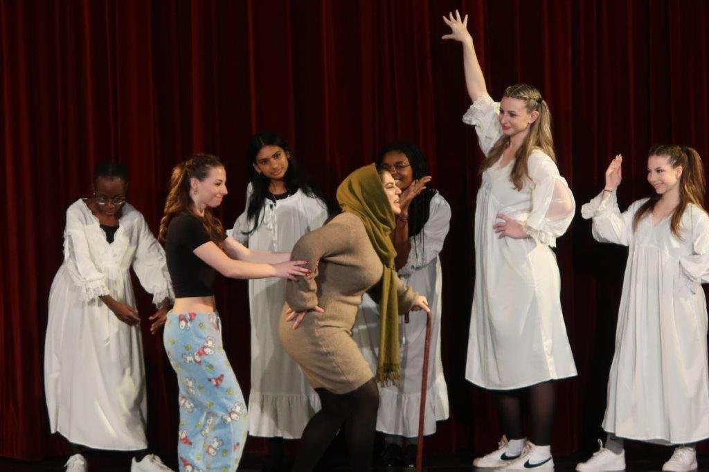  Actors dressed in white gowns in the rear as two actors walk in front; one in brown wearing a green headscarf and walking with a cane, and the other helping the former. 