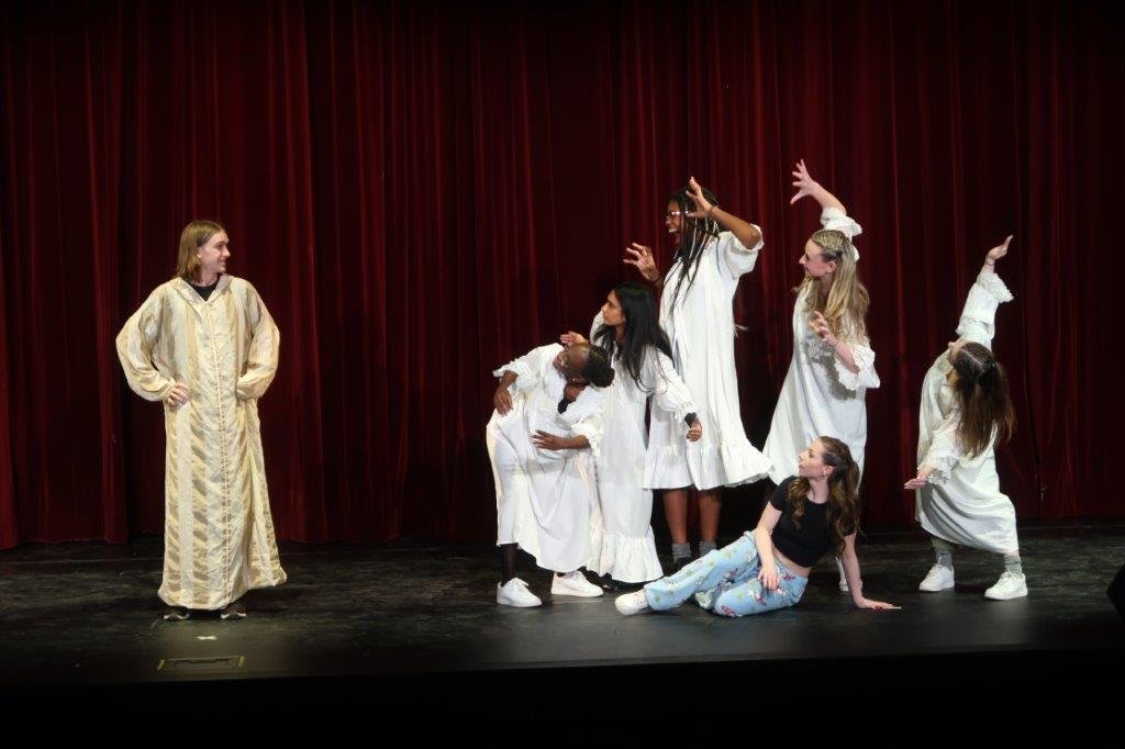  Actor on left in golden gown looks on at five actors in white gowns paused in scary poses looking back, and one actor laying on the floor below the five. 