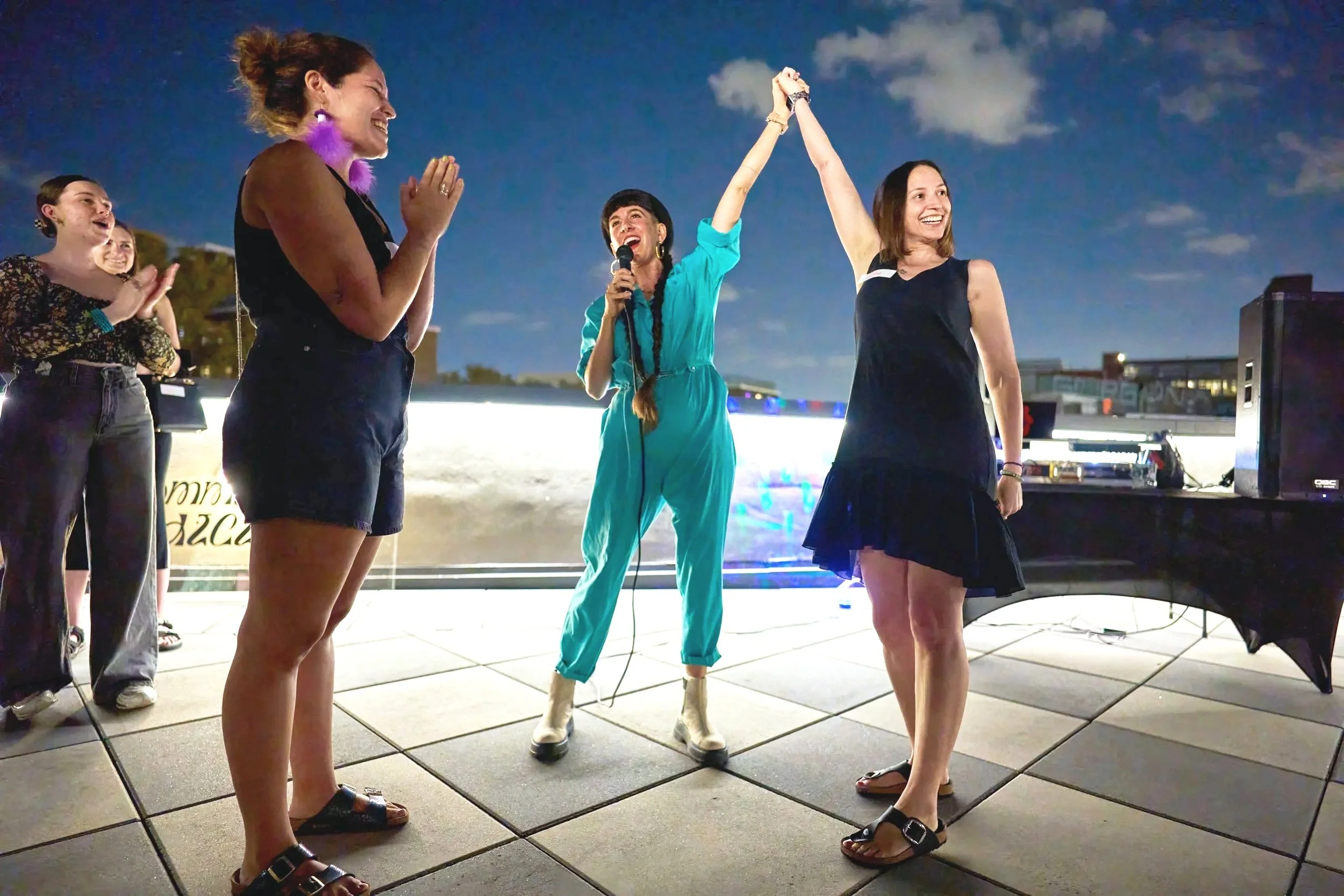 Two women practicing yoga indoors, sitting on yoga mats and smiling. One woman has red hair and wears a coral workout outfit. The other woman has dark hair, tied back, wearing a light green sports bra and leggings. A water bottle and a thermos are on the floor beside them, and a potted plant is in the background.