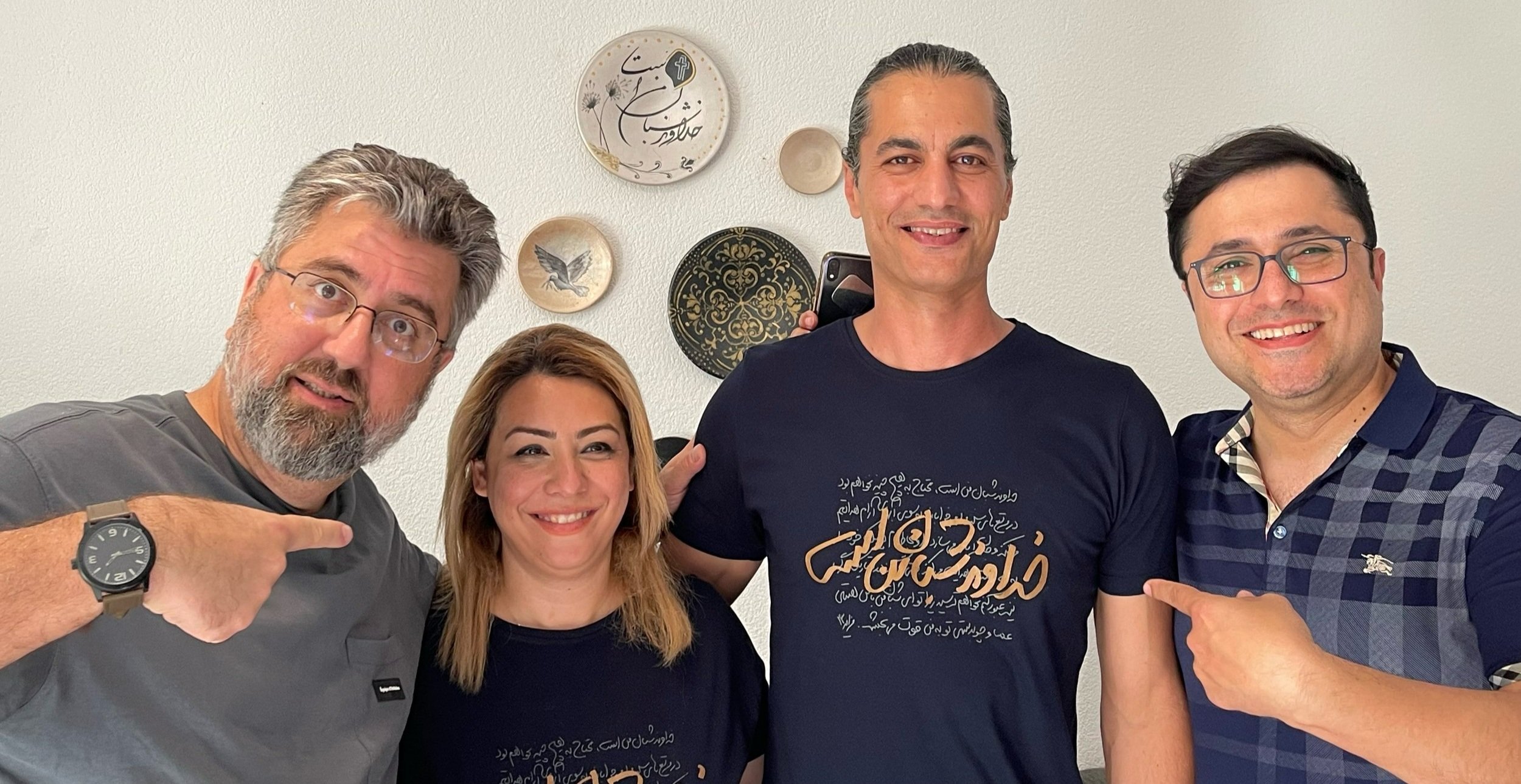   Asafeh and Rambod wearing Psalm 23 shirts with Psalm 23 in the background on wall-mounted plates. Surrounding them are Sasan and Dariush.  