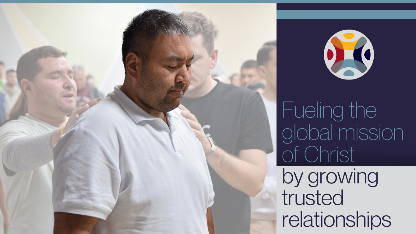 a man stands with two men behind him as they lay hands on his shoulders. They all have their eyes closed. Next to the image is a graphic with the words "fueling the global mission of Christ by growing trusted relationships."