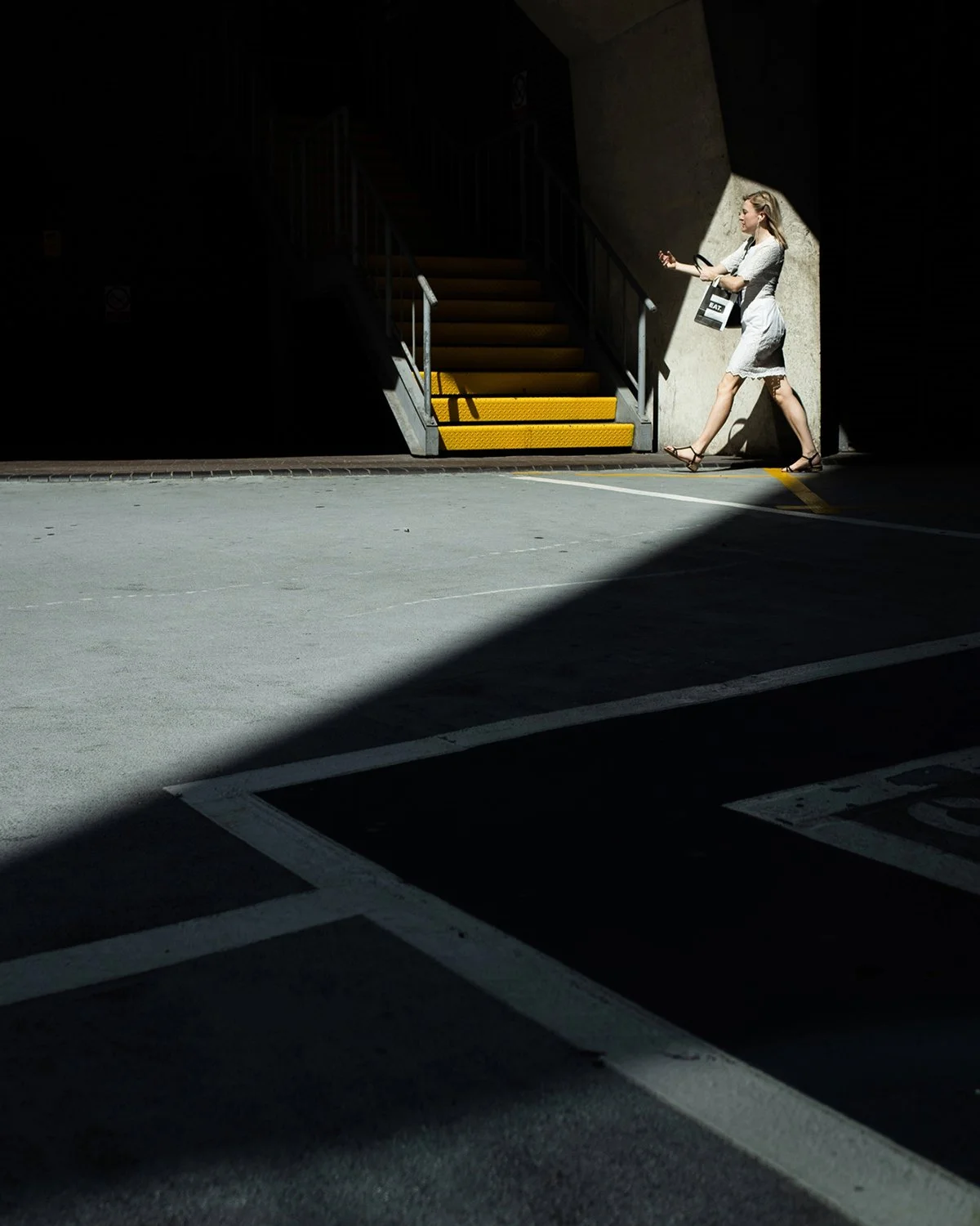 A woman in a white dress walking past a dark tunnel entrance with stairs and yellow safety strips, illuminated by sunlight.