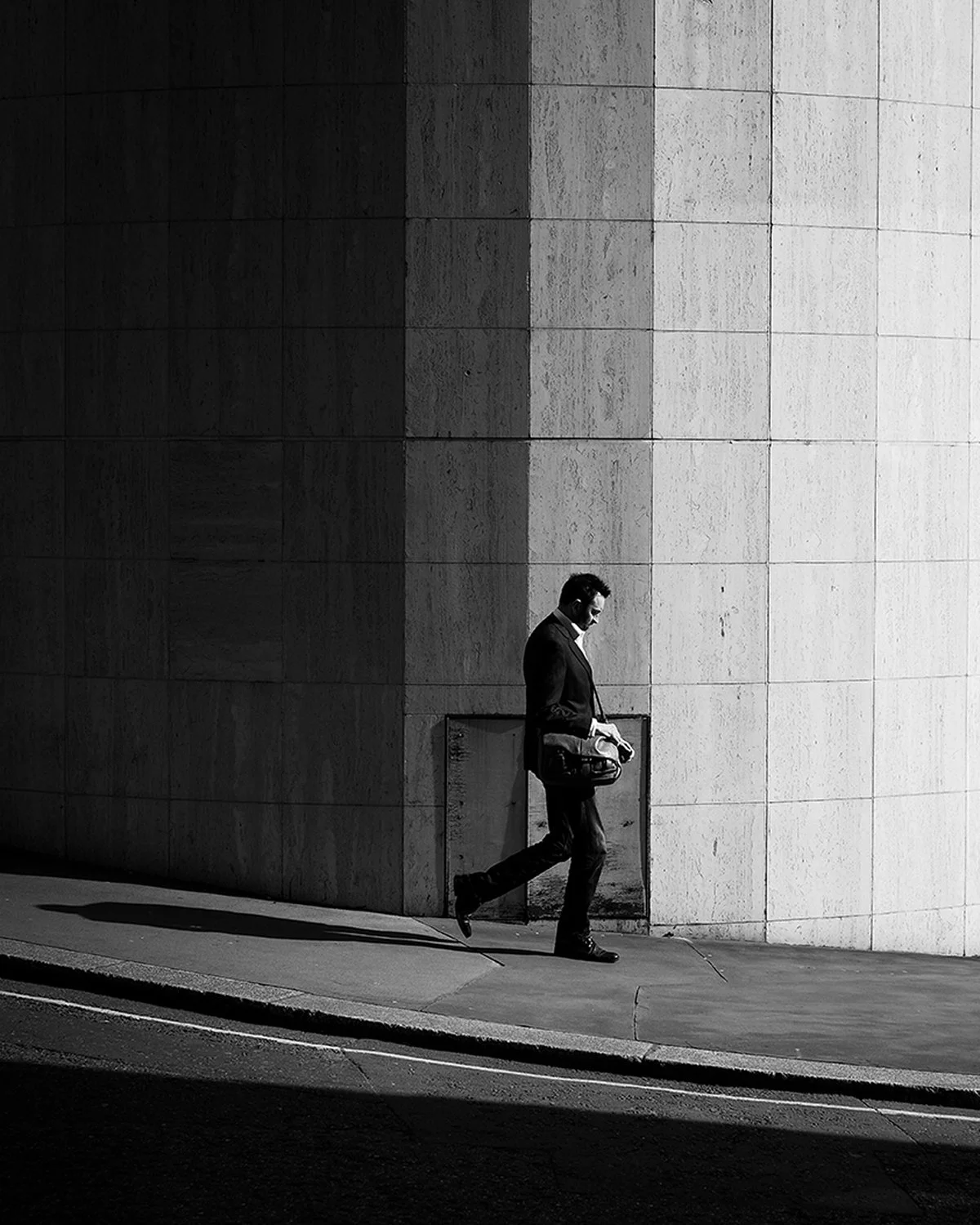 Black and white photo of a man in a suit walking on a city sidewalk, carrying a bag and holding a phone, with a large concrete wall in the background.
