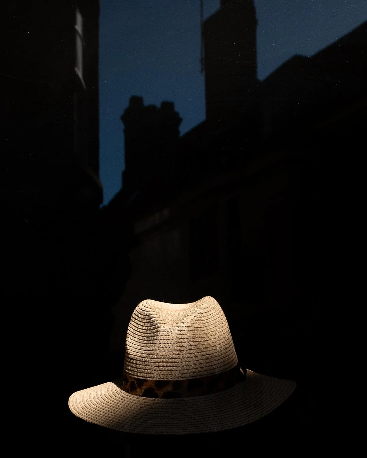 A straw hat with a leopard print ribbon on a dark reflective surface, with city buildings and a dark night sky in the background.