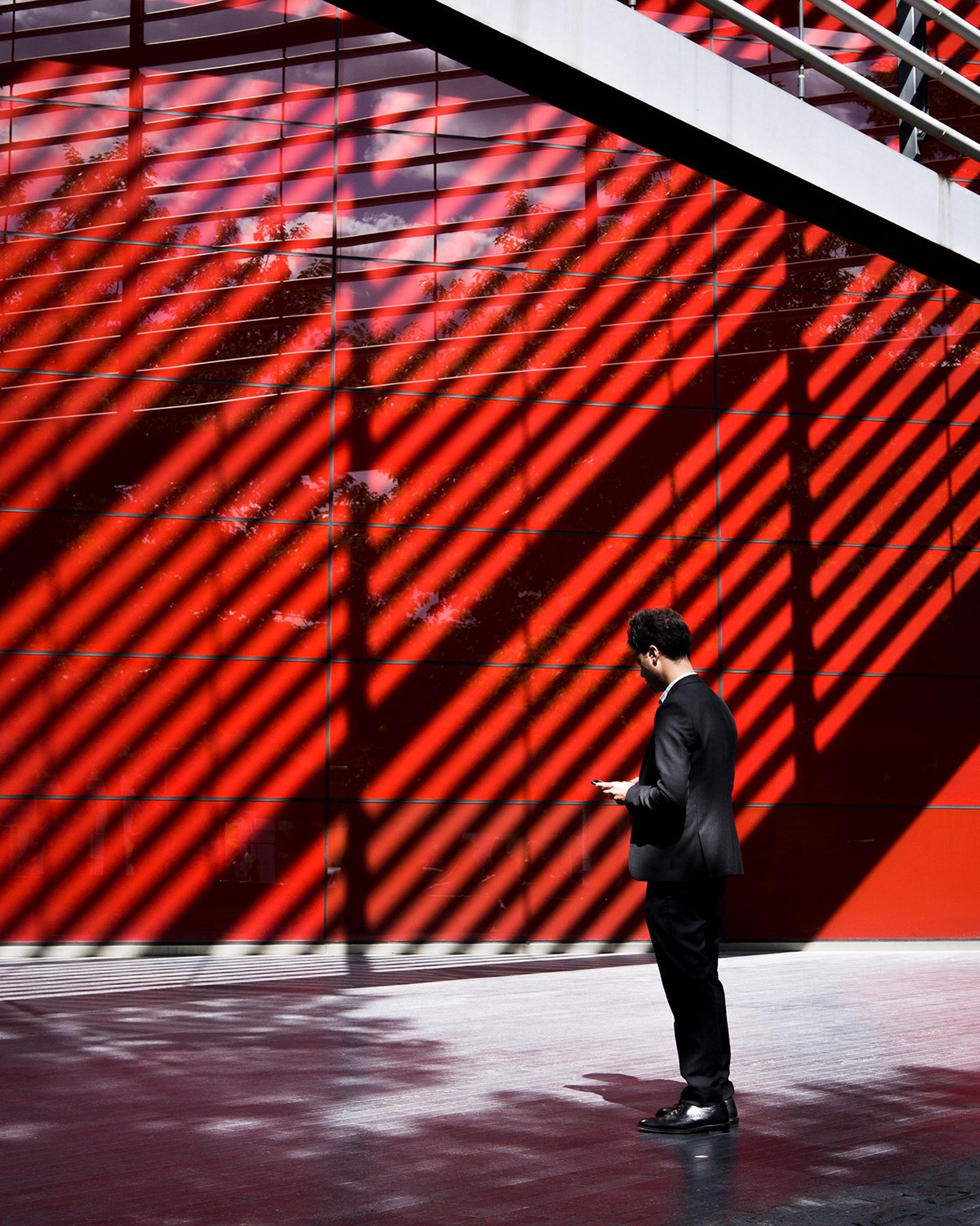 A man in a black suit looking at his phone in front of a red wall with diagonal shadows cast by a structure above.
