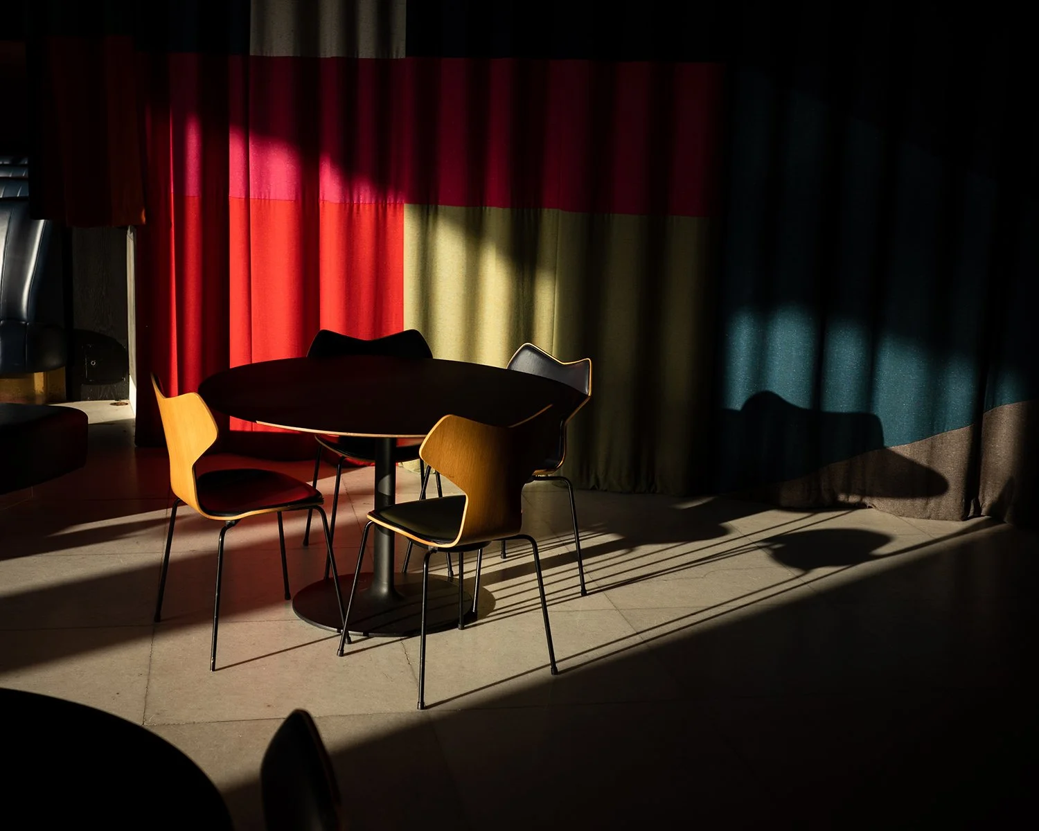 A round table with four chairs in a room, backlit by sunlight. The background has colorful curtains with red, green, and blue colors, creating shadows on the floor.